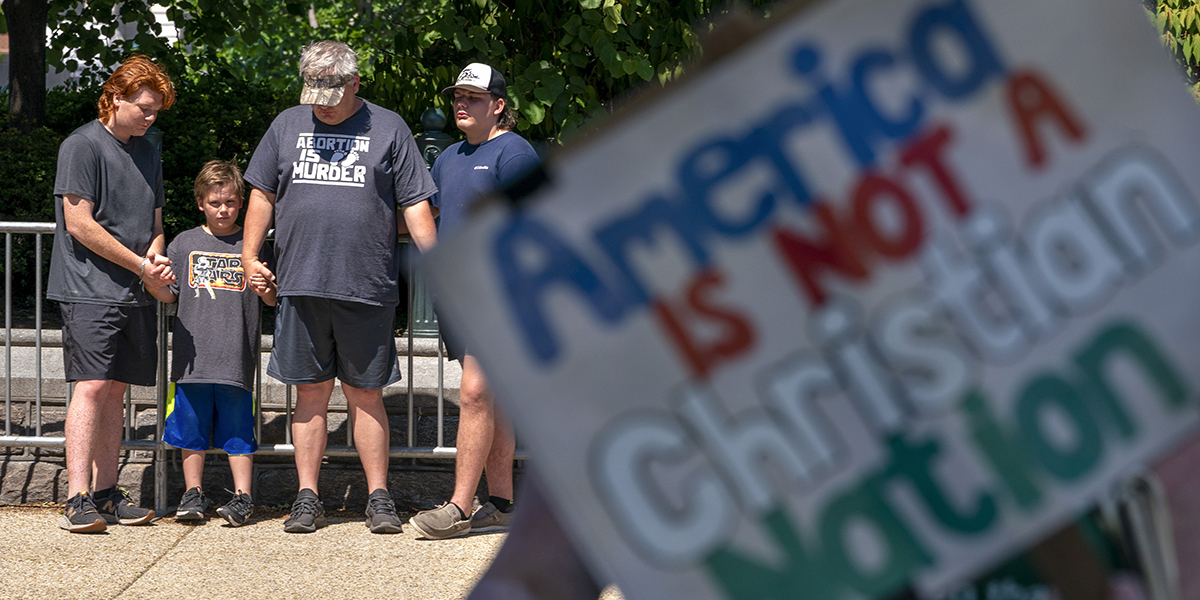 En far och hans söner från Florida har på sig T-shirt mot abort och ber, medan aborträttsanhängare protesterar utanför Högsta domstolen, med en skylt som säger "Amerika är inte en kristen nation", onsdagen den 29 juni 2022 , i Washington.