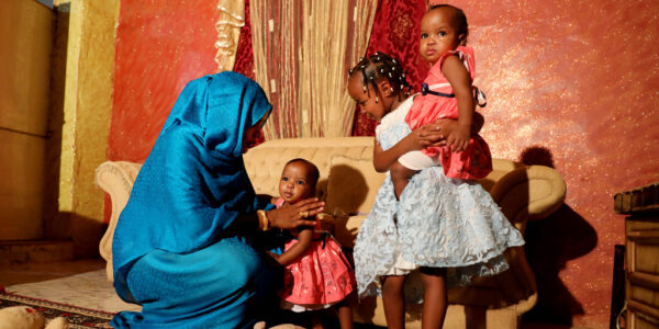 Kvinna i turkos klädedräkt sitter på huk, leker med flera små barn.