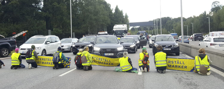 människor i varselvästar sitter framför bilar i protest