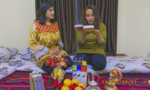 Två kvinnor sitter på golvet med frukter, bland annat granatäpple framför sig. Kvinnan till höger läser ur en bok.