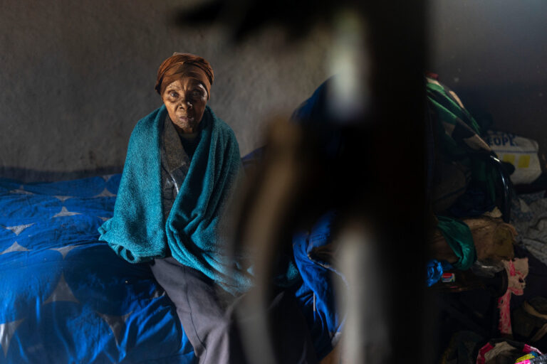 En kvinna sitter ner på en säng svept i en turkos filt och ser in i kameran.