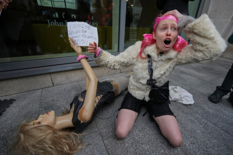 Kvinna sitter på marken bredvid en skyltdock och håller upp ett textat plakat.