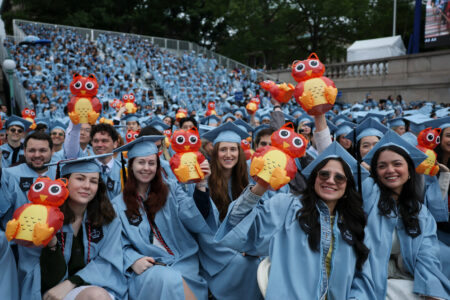 Studenter i ljusblå kappor och hattar håller upp små rödgula ugglor.