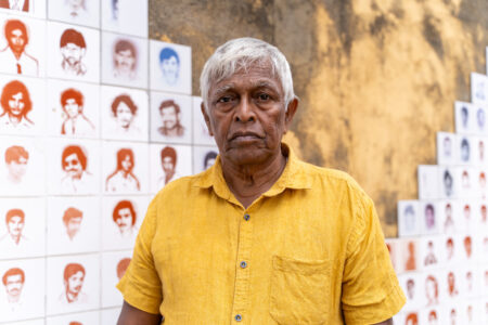 Brito Fernando i gul skjorta står framför vägg med porträtt av personer som utsatts för påtvingade försvinnanden.