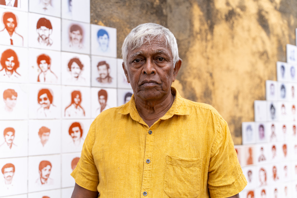 Brito Fernando i gul skjorta står framför vägg med porträtt av personer som utsatts för påtvingade försvinnanden.