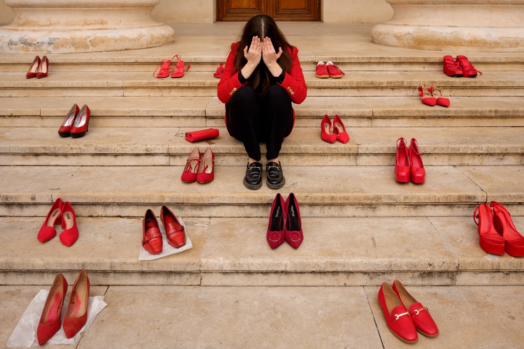 Röda skor uppställda på en bred trappa, kvinna med röd kavaj sitter ner, håller händerna för sina ögon.