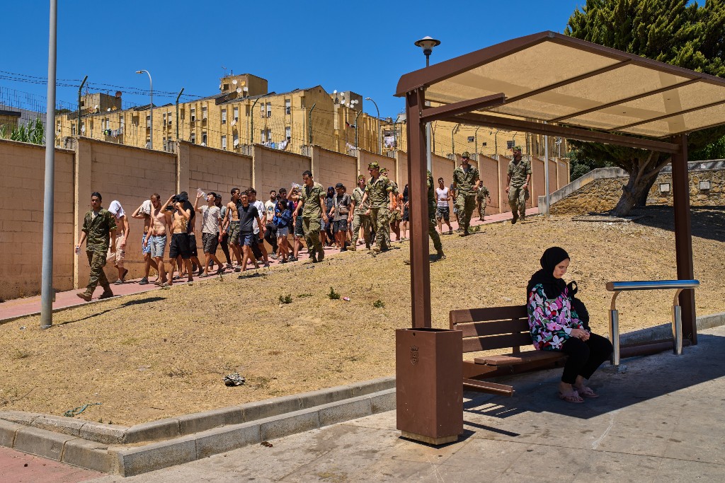 Migranter i Ceuta förs till gränsen till Marocko av spanska soldater, i förgrunden en kvinna som väntar vid en busshållplats.